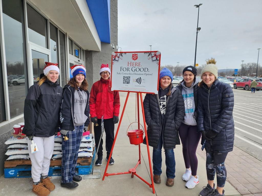 National Honor Society students and advisor wearing winter coats and hats, outside of a store, ringing the bell for Salvation Army. They are standing in a line, next to the red Salvation Army kettle.