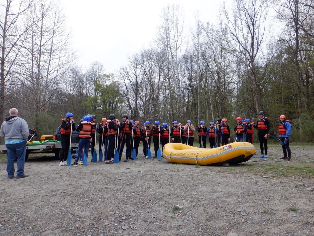 Students stand in a line with life vests on, holding oars. They are standing in front of a raft