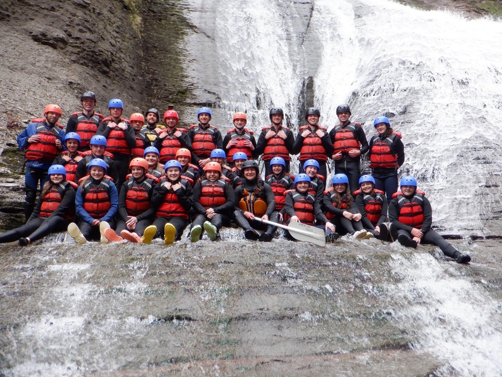 Students pose in front of a waterfall, some are standing, some are sitting. They are wearing life vests and helmets